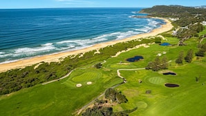 Vista aérea de un campo de golf costero con playa, búnkeres de arena y exuberante vegetación.