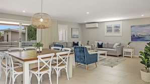 Una sala de estar moderna con una mesa de comedor de madera, sillas blancas, un sofá azul y un sofá gris. Hay un ventanal con persianas, un equipo de aire acondicionado en la pared y un cuadro sobre el sofá.