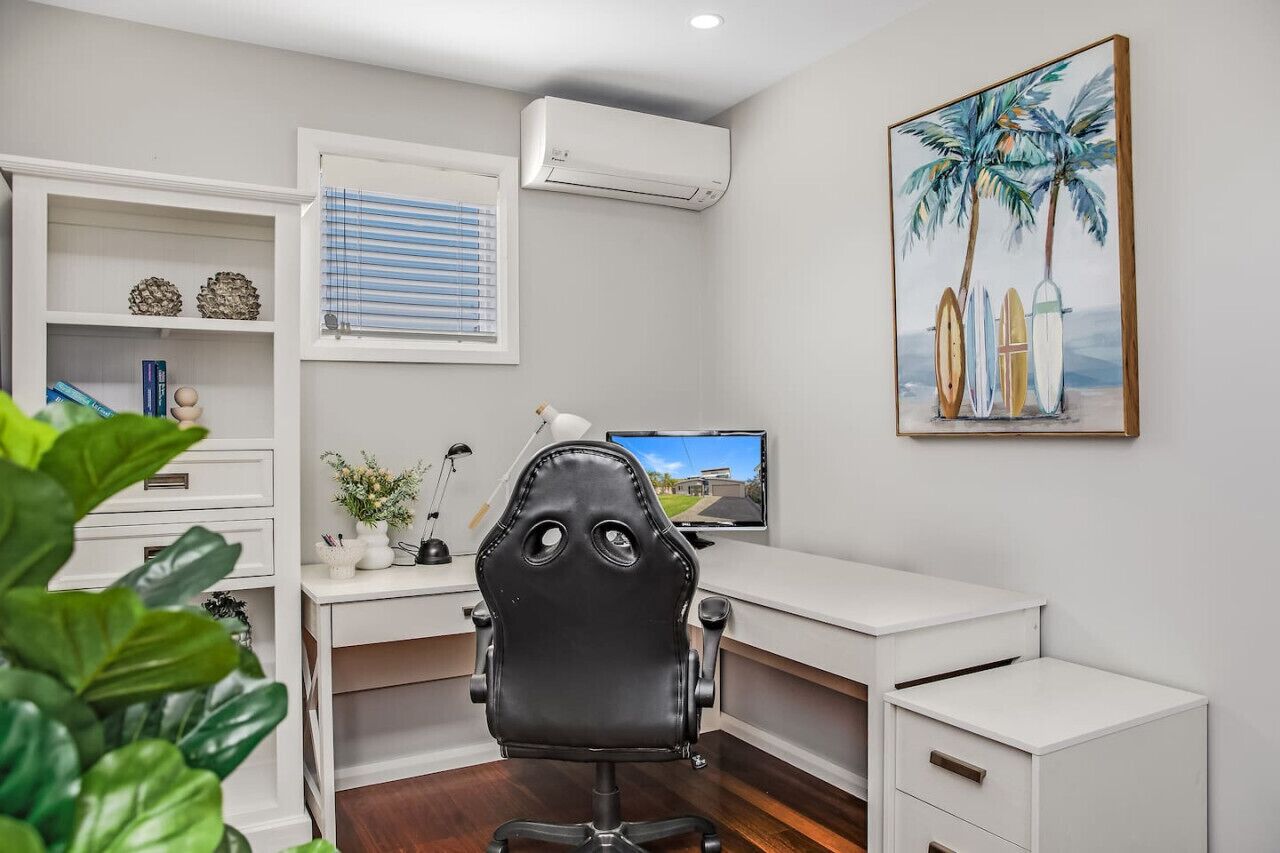 A modern office with a white desk, a black office chair, a computer, and a window with blinds.