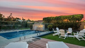 Una piscina con cascada, rodeada por una terraza de madera y sillas reclinables blancas.