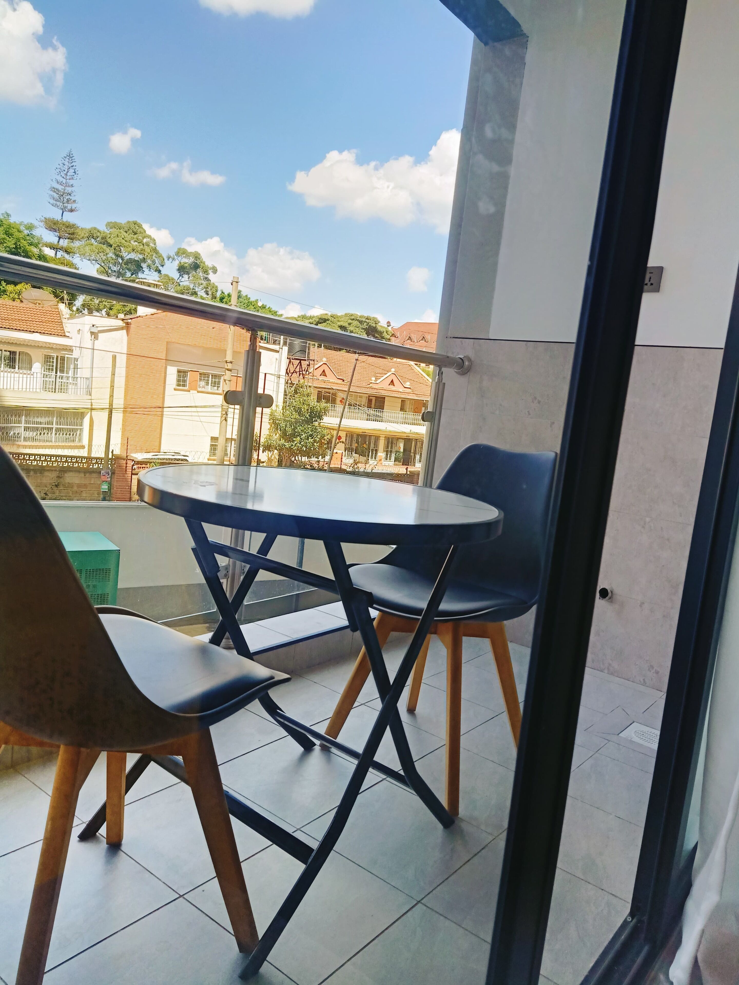 A balcony with a round table and two chairs, a green air conditioning unit, and a view of buildings and trees.