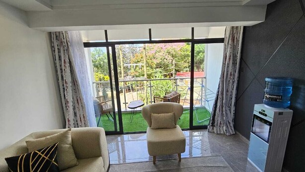 A modern living room with a sofa, armchair, and a glass coffee table. A water dispenser is visible on the right. - Lux Suites Gatundu Heights Kileleshwa (Nairobi)