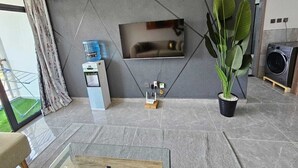 A modern living room with a flat-screen TV mounted on a geometric wall, a water dispenser, a potted plant, and a washing machine in the background. - Lux Suites Gatundu Heights Kileleshwa (Nairobi)