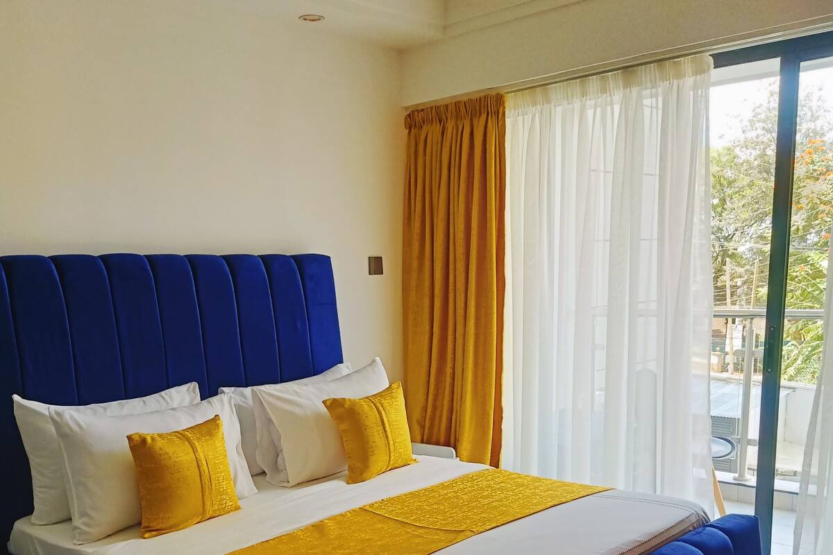 A modern bedroom with a blue upholstered headboard, a large bed with white and yellow bedding, and a view of the outdoors through a large window with sheer curtains.