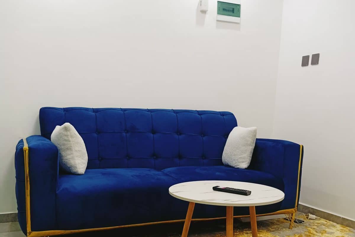 A blue sofa with two white cushions, a small white table with a remote control, and a patterned rug on a light-colored floor.