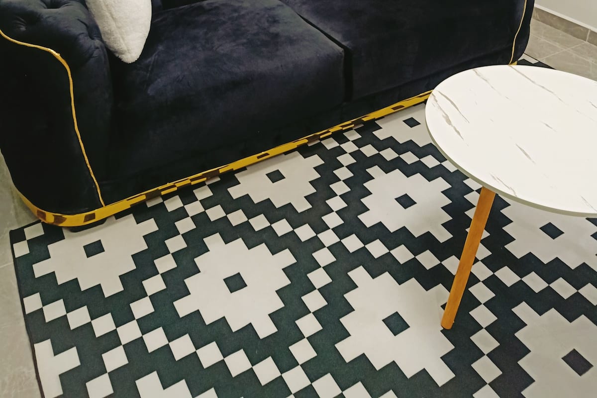 A dark blue tufted sofa with two white cushions, a patterned rug, and a small white round table with wooden legs.