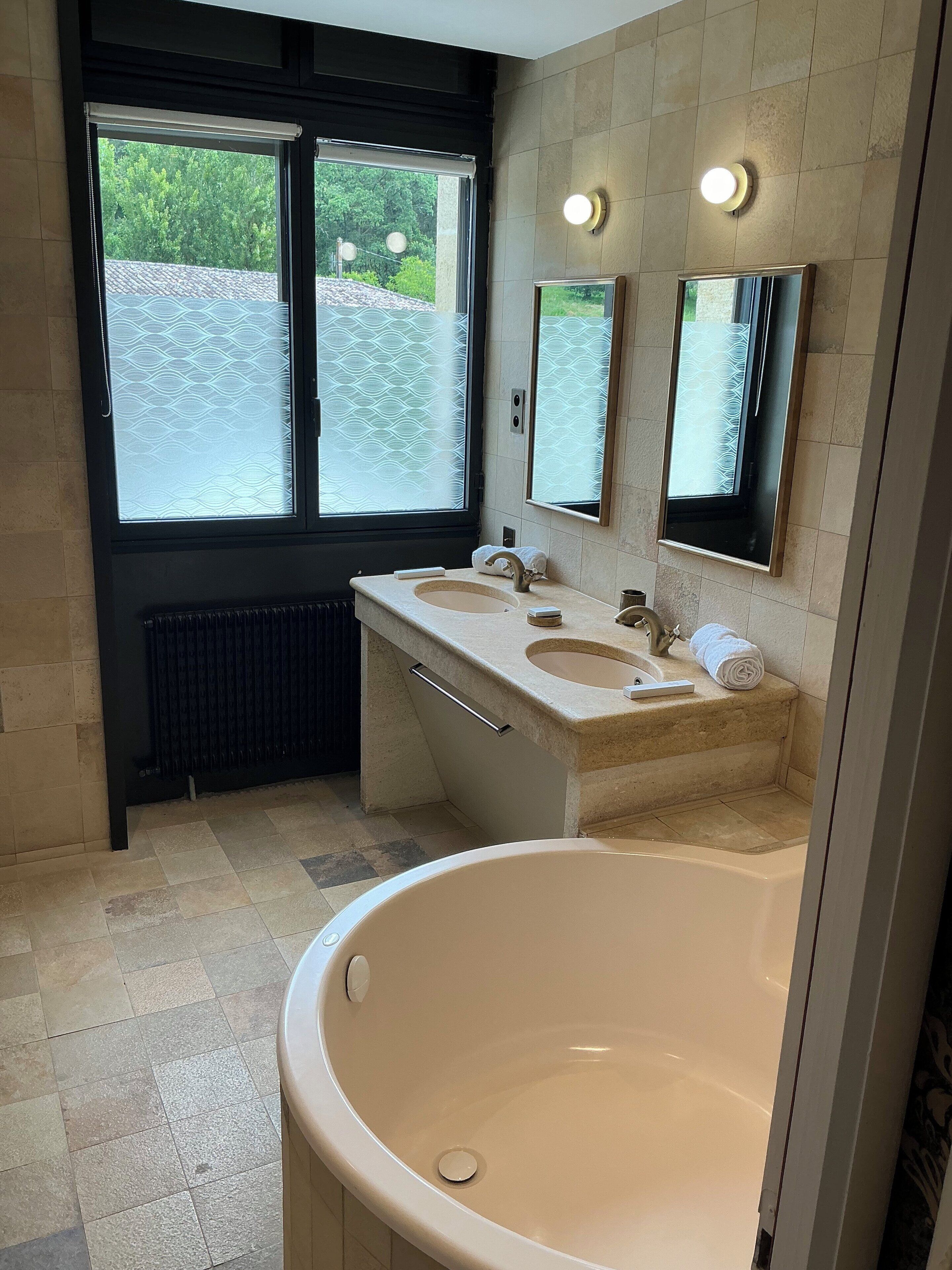 Un baño moderno con dos lavamanos, una bañera grande y un ventanal con vistas a la vegetación.