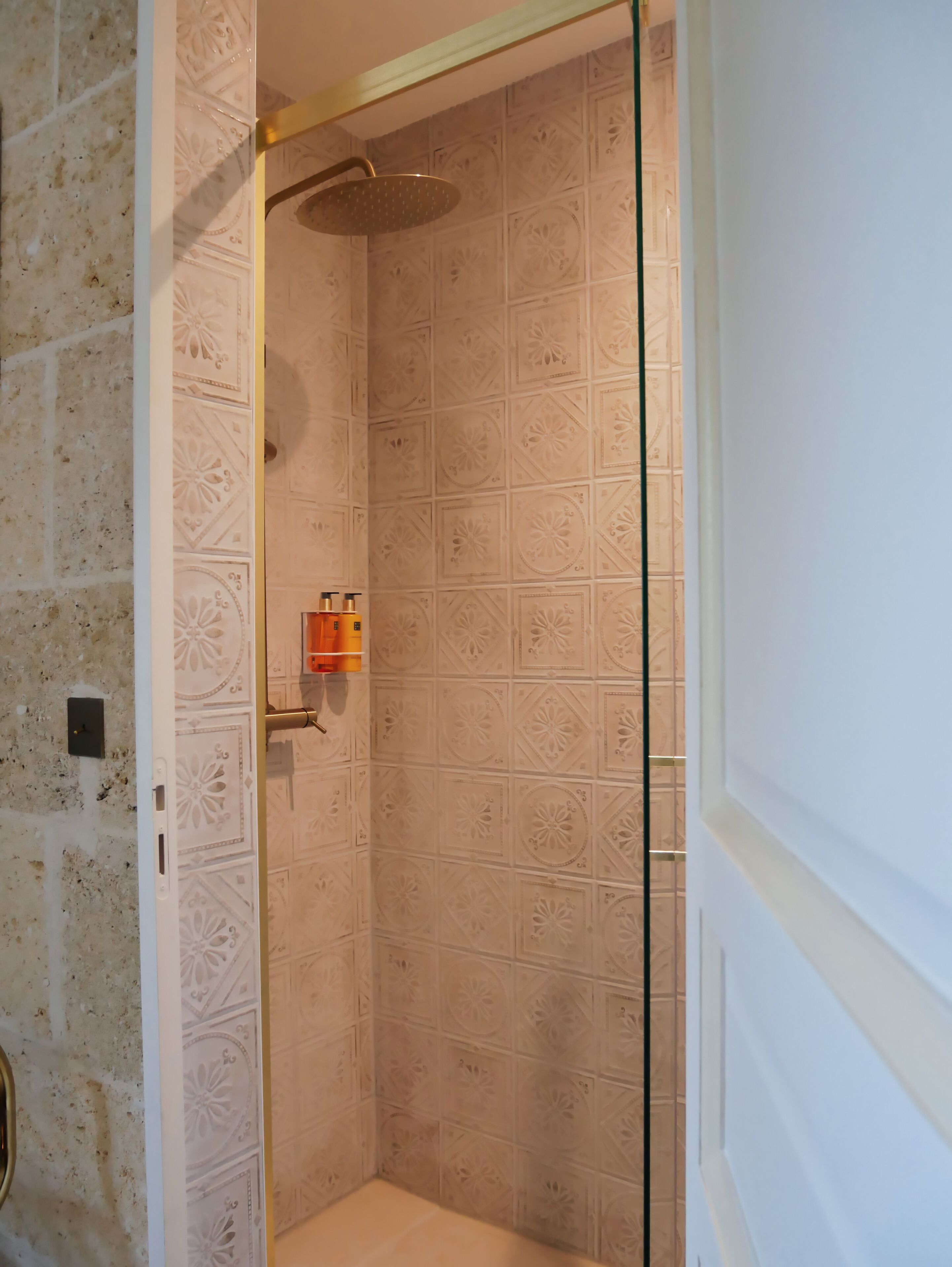 A shower with a glass door, a wall with a geometric tile pattern, and a small shelf with toiletries.
