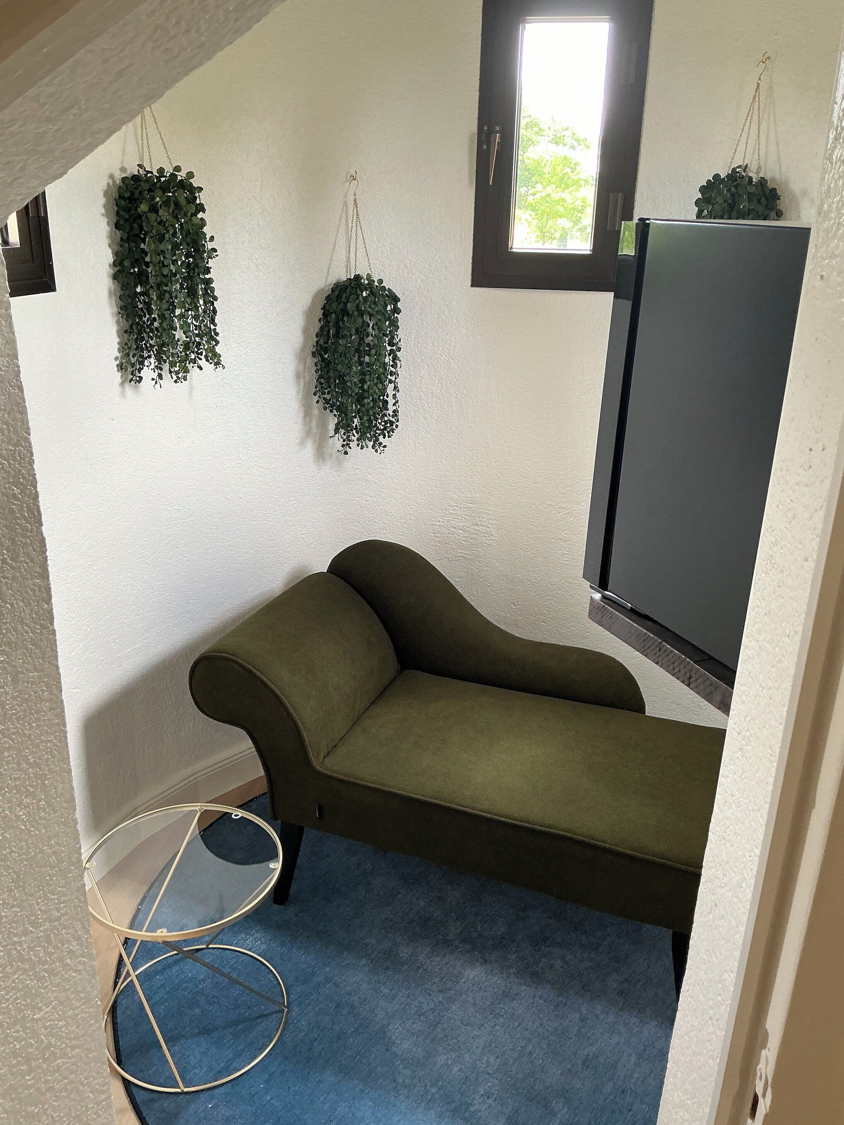 Une pièce d’angle avec un canapé vert, une petite fenêtre et deux plantes suspendues.