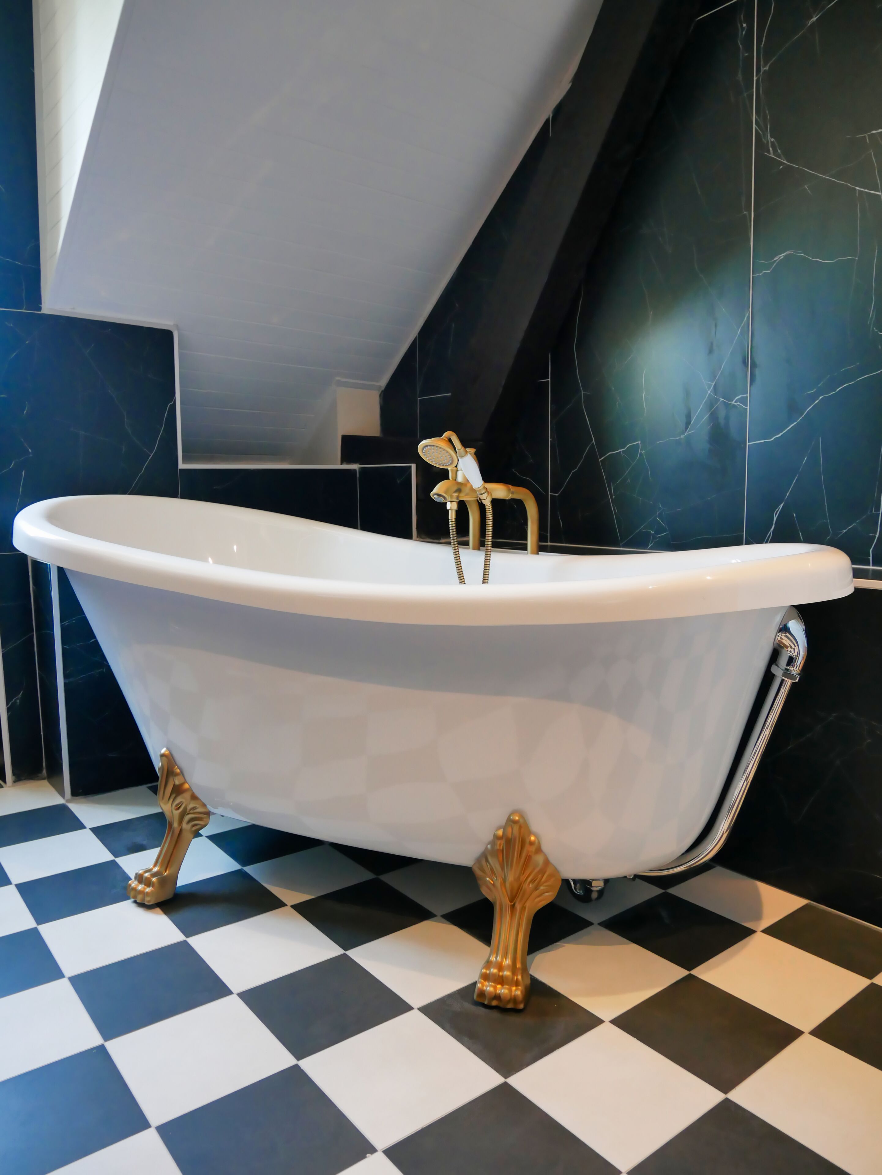 Een vrijstaand bad met klauwvoeten in een badkamer met zwart-wit geruite vloer en donkere marmeren wanden.