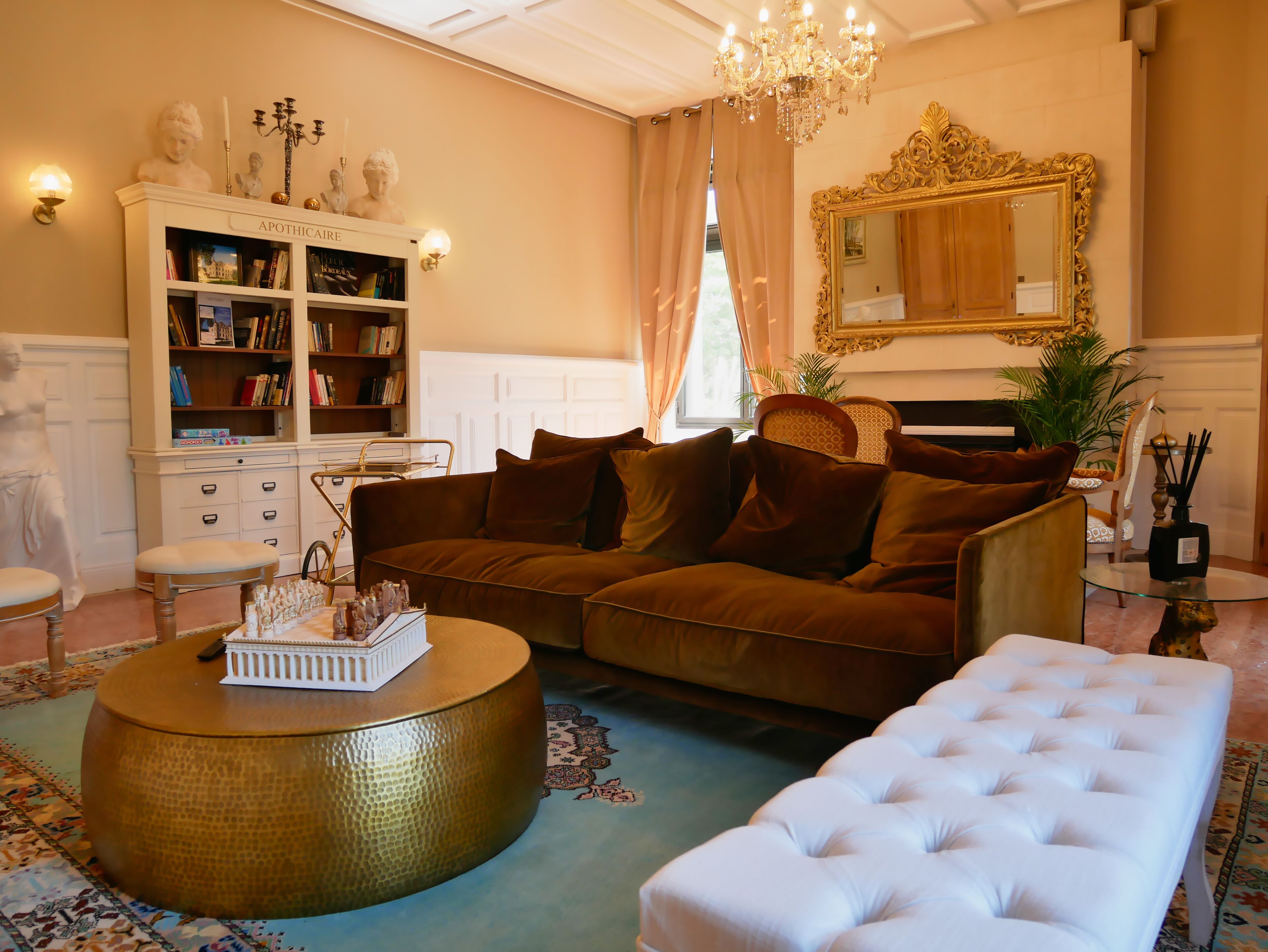 A living room with a brown sofa, a white ottoman, a round coffee table, a bookshelf, and a chandelier.