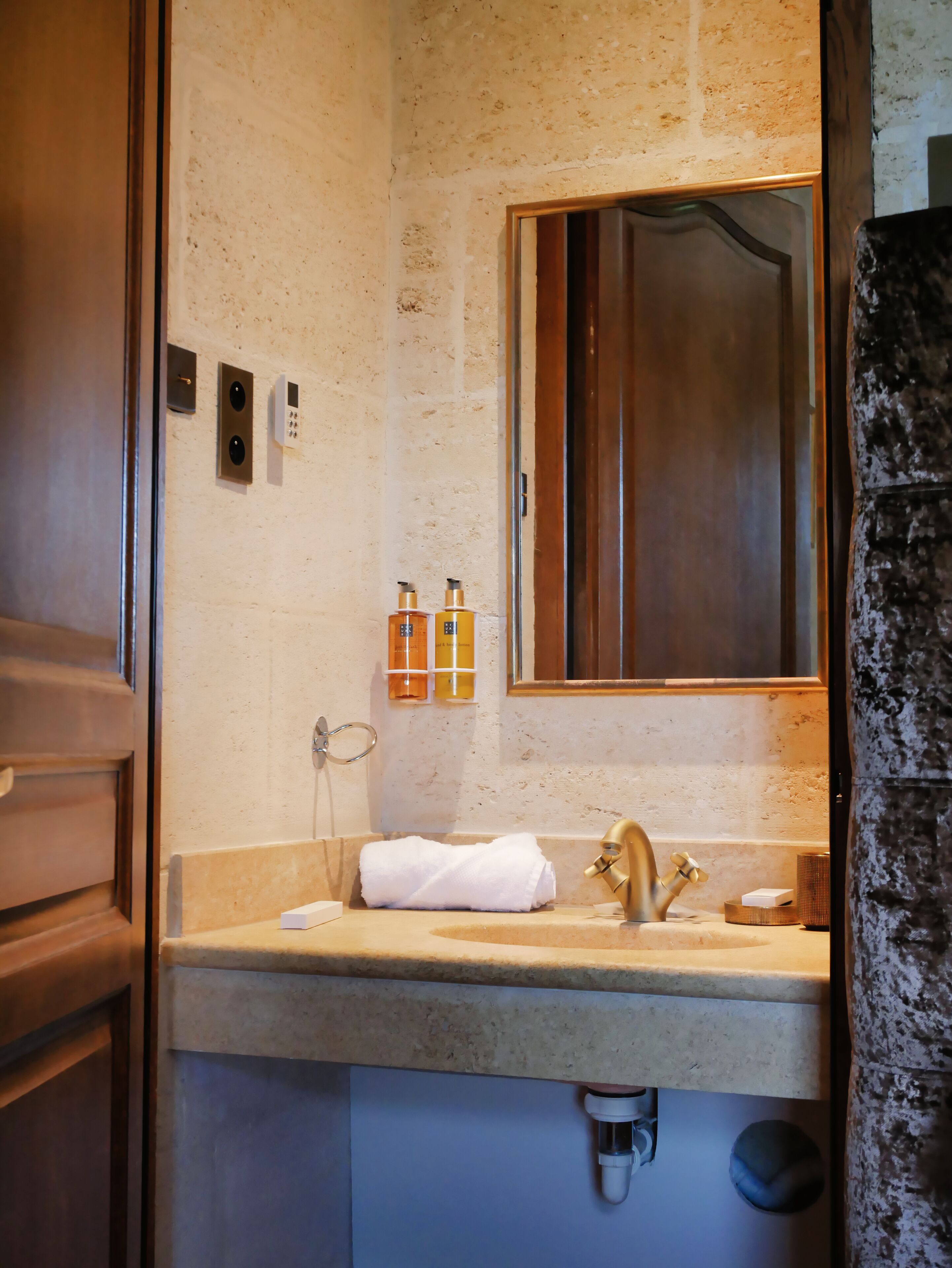 A bathroom with a wooden vanity, a sink with a gold faucet, a towel, and two bottles of toiletries on the wall.