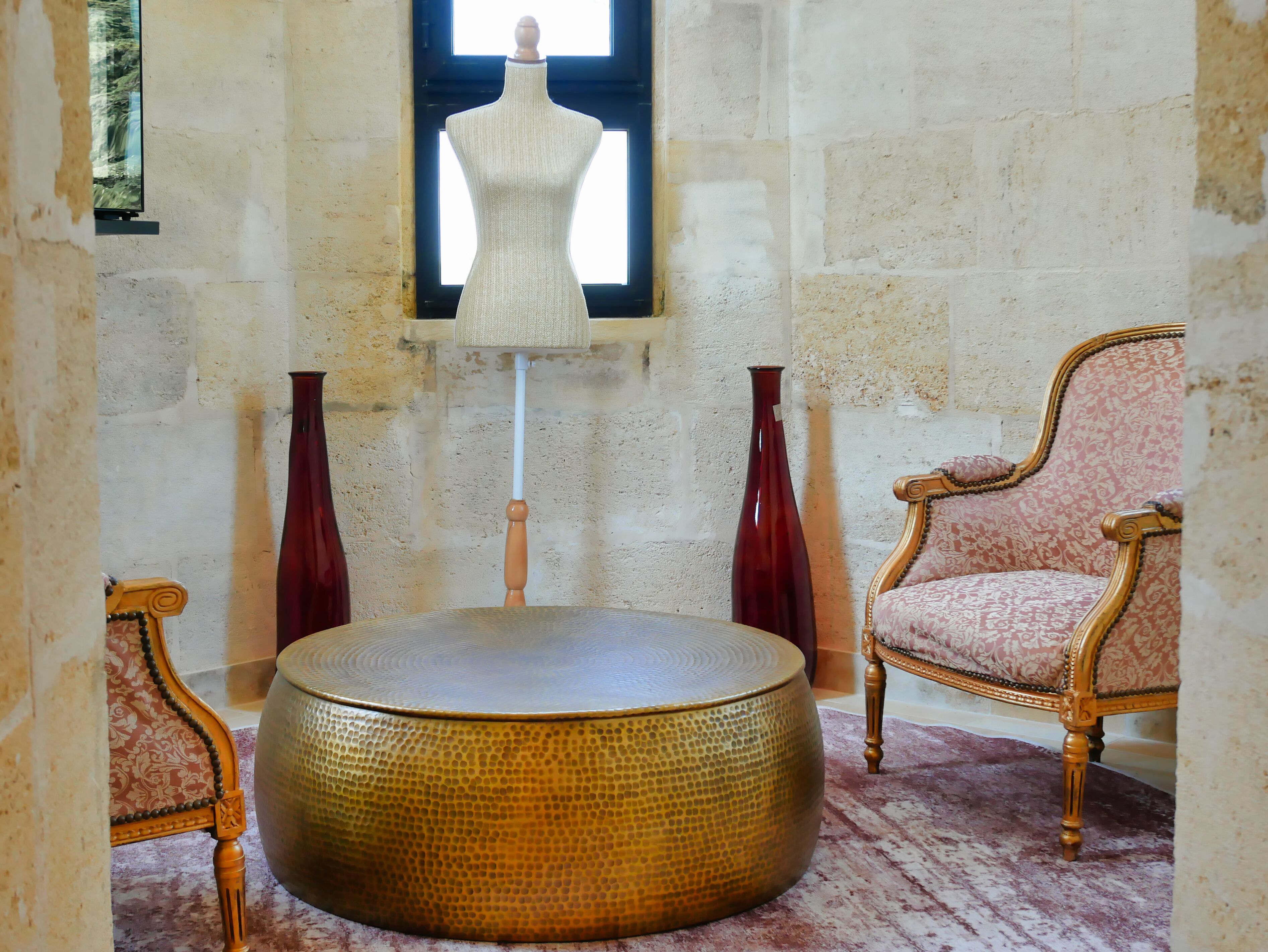 Ein Zimmer mit einem runden, strukturierten Ottomanen, einem Vintage-Sessel, einer Schaufensterpuppe und einer hohen Vase.