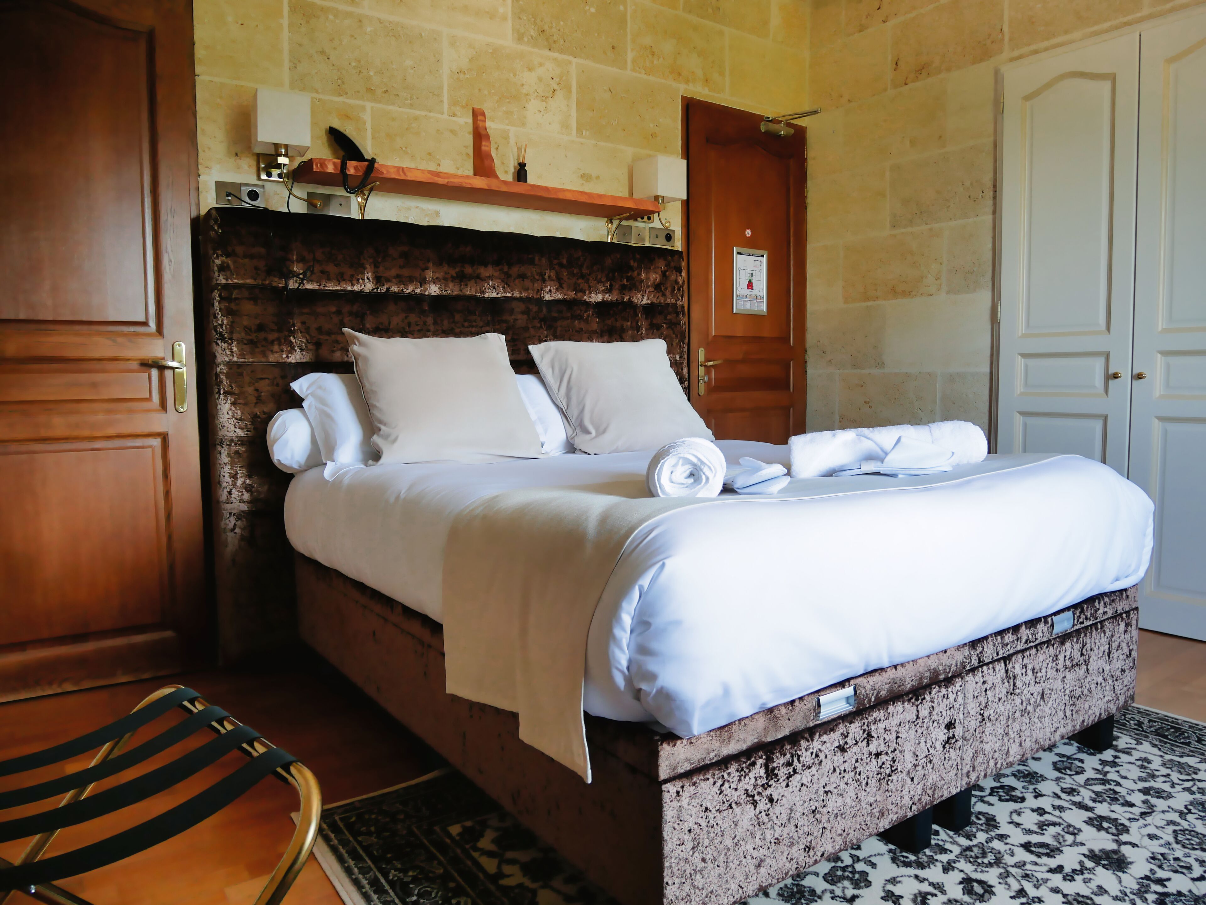 A neatly made bed with white linens and pillows, a wooden headboard, a small shelf above the bed, and a patterned rug on the floor.