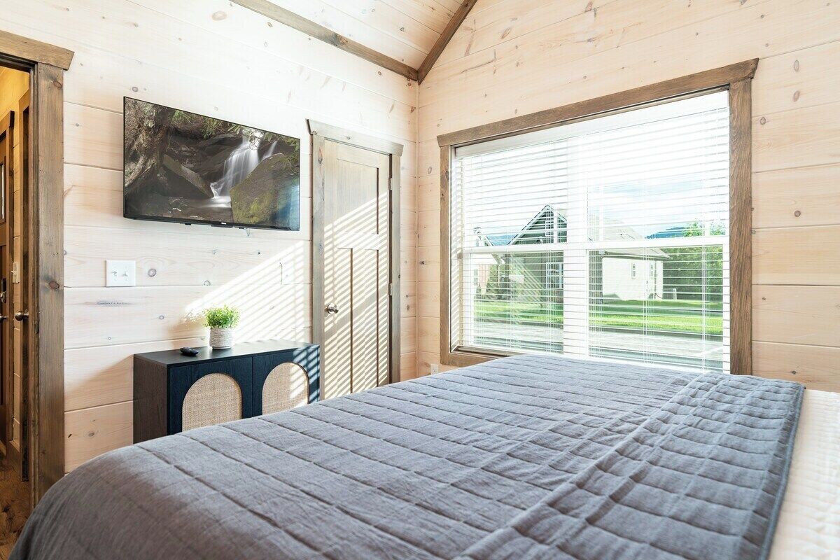 A bedroom with a bed, a TV mounted on the wall, a window with blinds, and a small table with a plant.
