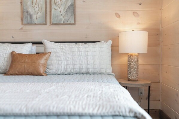 A neatly made bed with a white quilt and two pillows, a wooden wall with two framed botanical prints, and a bedside lamp on a small table.