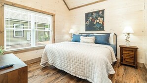 Un dormitorio con piso de madera, una cama con colcha blanca acolchada, dos lámparas de noche, un cuadro en la pared y una ventana con persianas.