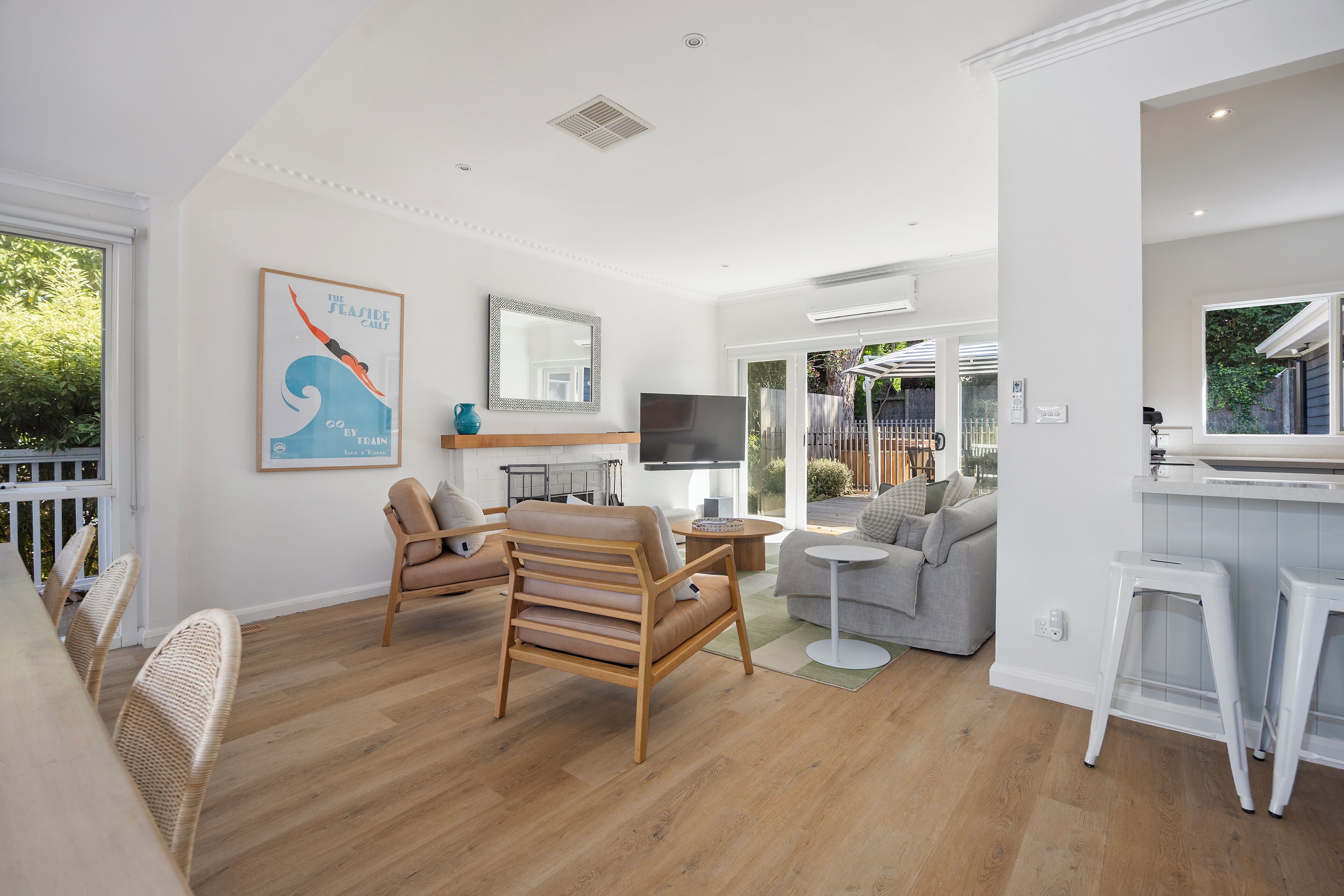 Una sala de estar moderna con piso de madera, un sofá gris, una silla de madera, un televisor y una zona de cocina con encimera blanca y taburetes.