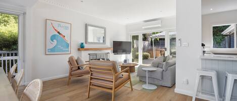 Una sala de estar moderna con piso de madera, un sofá gris, una silla de madera, un televisor y una zona de cocina con encimera blanca y taburetes.