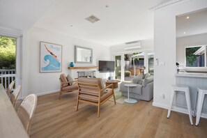 Uma sala de estar moderna com piso de madeira, um sofá cinza, uma cadeira de madeira, uma TV e uma área de cozinha com bancada branca e banquetas.