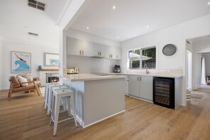 A modern kitchen with a central island, white cabinetry, and a built-in dishwasher. There are bar stools, a coffee machine, and a clock on the wall. - Portsea Beach Haven with Hot Tub (Portsea)