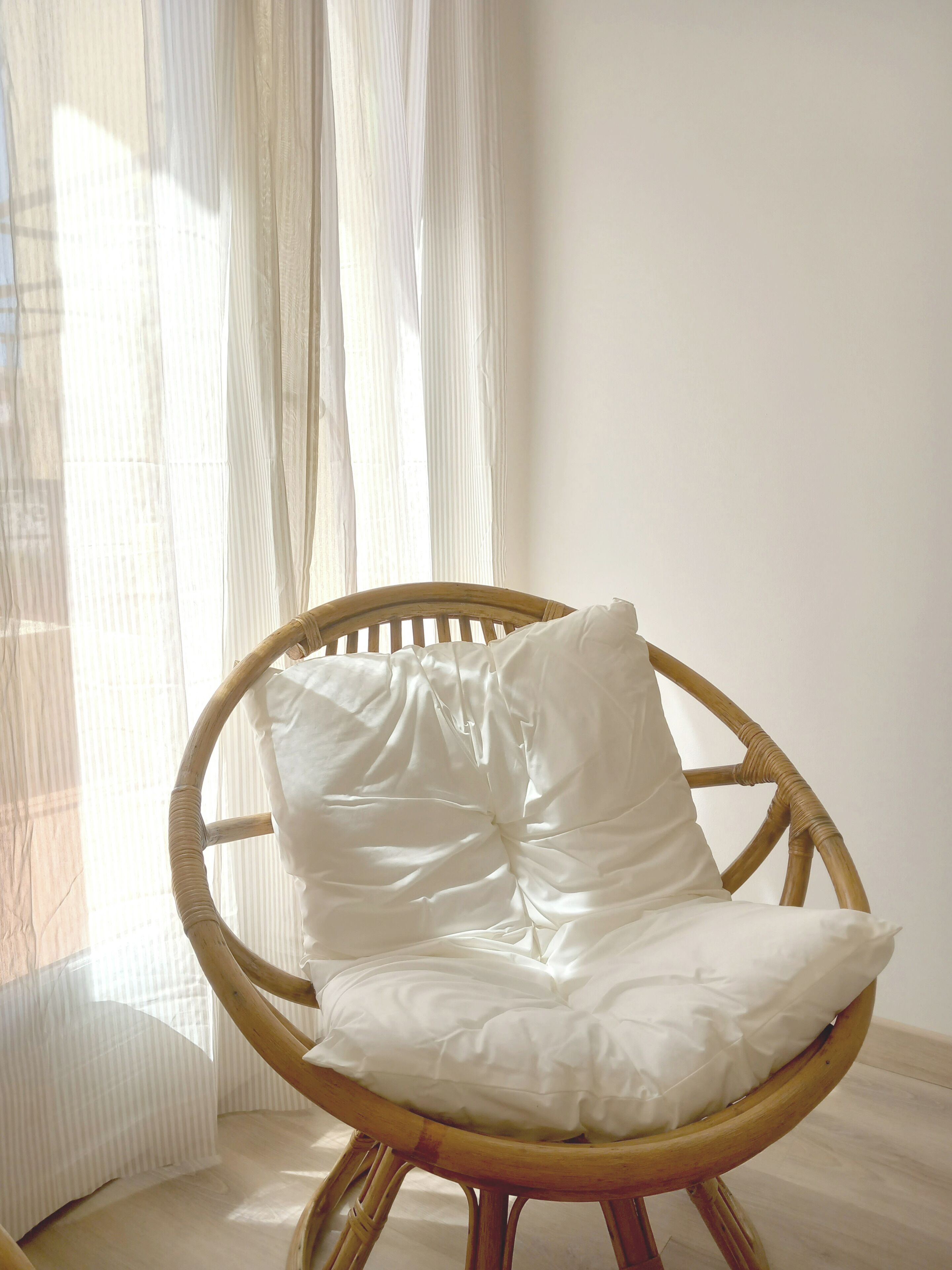 A wicker chair with white cushions in a room with sheer curtains and a light-colored floor.