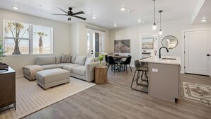A spacious living area with a sofa, ottoman, and round coffee table, a dining area with a table and chairs, a kitchen with a sink and island, and a ceiling fan.