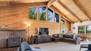 A spacious living room with a fireplace, wooden walls, and large windows offering views of trees. - The Bodega House~Bodega Bay~Near Beach~Family Fun~Game Room~Sonoma Coast Fun! (Bodega Bay)