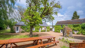 A rustic outdoor seating area with a wooden picnic table and benches, surrounded by stone buildings with ivy-covered walls and a grassy lawn. - Propriété au Calme Avec Piscine au Coeur de L'anjou ! Idéale Pour les Familles (Baugé en Anjou)
