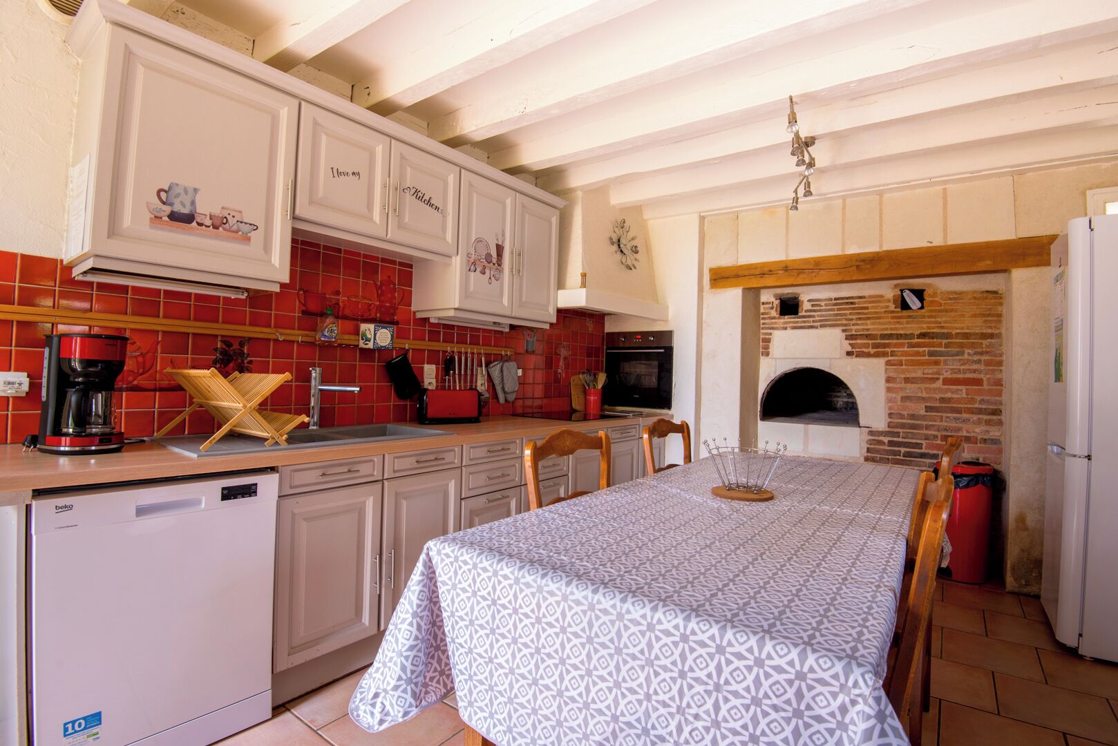 A kitchen with white cabinets, a red tiled backsplash, a built-in oven, and a dining table with a patterned tablecloth.