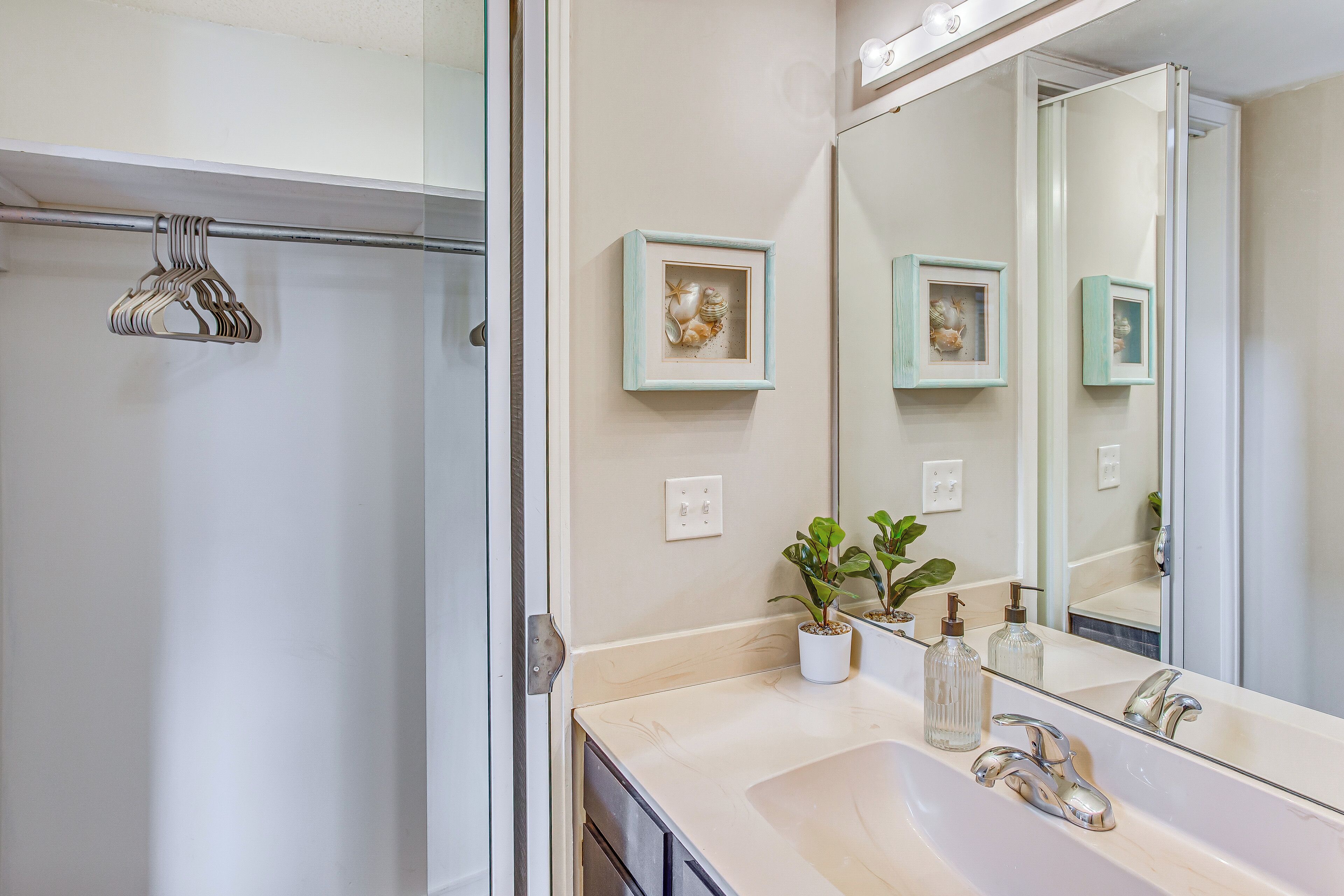 Une salle de bain avec un grand miroir, deux tableaux encadrés, un lavabo avec deux robinets et un espace douche.