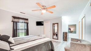 A bedroom with a bed, a ceiling fan, a TV, a chair, and a door leading to another room.