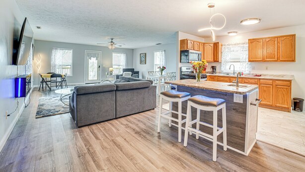 A modern kitchen with wooden cabinets, a central island with stools, and a dining area with a sofa and table.