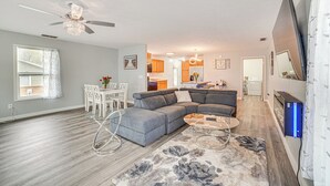 A modern living room with a sectional sofa, a dining area with a table and chairs, a kitchen in the background, and a ceiling fan.
