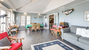 Een ruime woonkamer met een grijze bank, rode en houten stoelen, een eethoek met een groene tafel, en een plafondventilator.