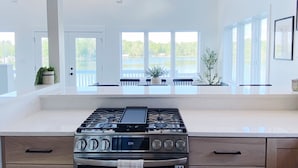 A kitchen with a gas stove, oven, and a large island with a sink. There are two pendant lights above the island, and large windows with a view of a lake. - [New!] Luxurious Lake Front Getaway!  (Wawaka)
