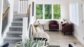 A spacious living room with a staircase, a chandelier, and large windows offering a view of greenery. - [New!] Luxurious Lake Front Getaway!  (Wawaka)