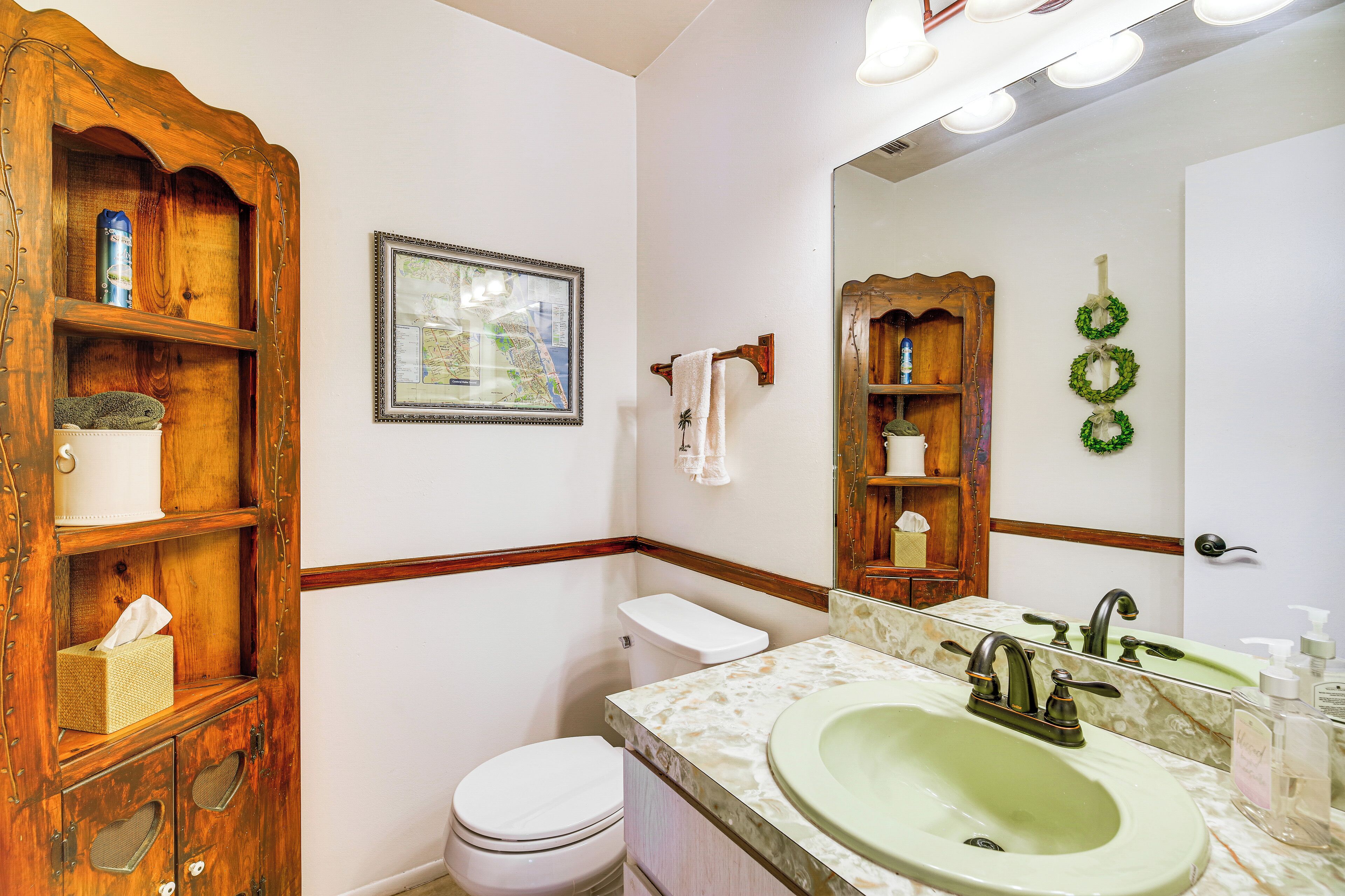 A bathroom with a wooden vanity, a white toilet, a green sink, and a mirror with lights.