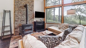 A cozy living room with a stone wall, a fireplace, a sofa, and a large window with a view of trees. - Luxe Appalachian Mountain Cabin w/ Hot Tub, Views (Warrensville)