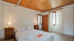 Un dormitorio con techo de madera, una cama con ropa de cama blanca, dos mesitas de noche con lámparas y una ventana con cortinas.