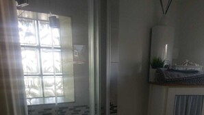 Et brusebad med glasvægge og flisebeklædt væg, et hvidt skab med glashylde og et lille trin til brusebadet.