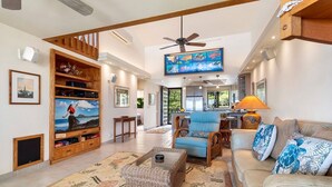 A spacious living room with a large TV, wooden entertainment unit, and a cozy seating area with a blue armchair and a sofa.
