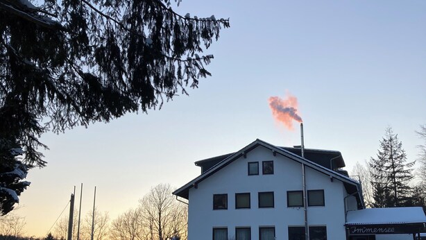 A building with a chimney emitting smoke, surrounded by snow-covered ground and trees. - Immensee III Apartment with lake view surrounded by forest (Ronshausen)