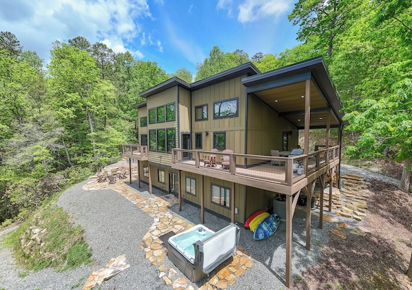 A two-story building with a deck, hot tub, and outdoor seating area surrounded by trees.