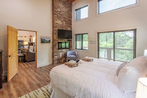 A bedroom with a large bed, a stone fireplace, a TV, and a view of the outdoors.