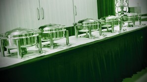 Ein Büfett mit mehreren abgedeckten Servierstationen auf einer langen Tafel, vor einem Hintergrund aus grünem Vorhang und einer Zierleiste.
