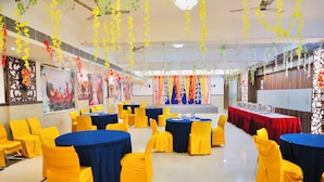Ein Festsaal mit runden Tischen, bedeckt mit blauen Tischdecken, und gelben Stühlen, verziert mit hängenden Blumenarrangements.