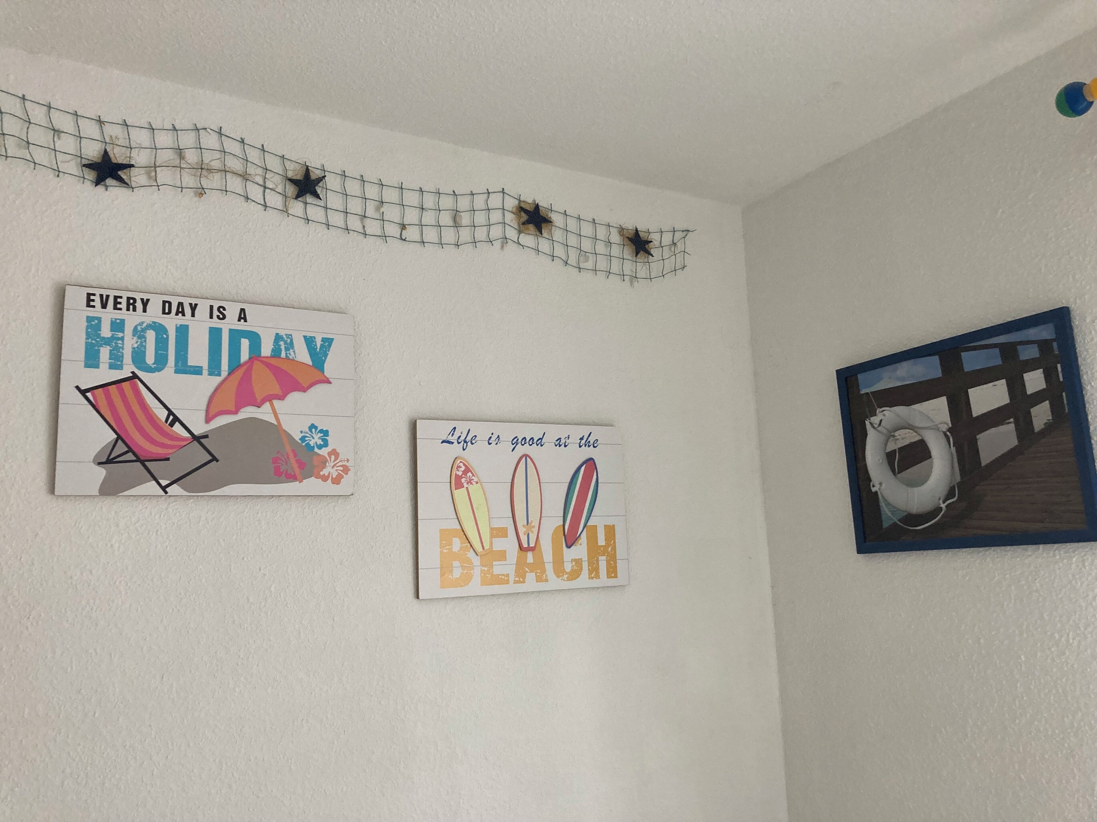 Three wall decorations: a beach chair with an umbrella, a surfboard with 'Life is good at the beach' text, and a lifebuoy on a wooden dock.