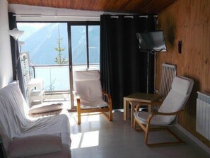 A room with a wooden ceiling, a sofa, two chairs, a small table, a TV, and a view of mountains through a large window.