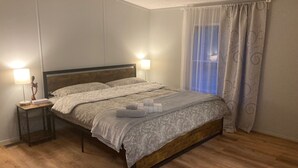 Una cama bien hecha con estructura de madera, dos lámparas de noche y una ventana con cortinas traslúcidas.